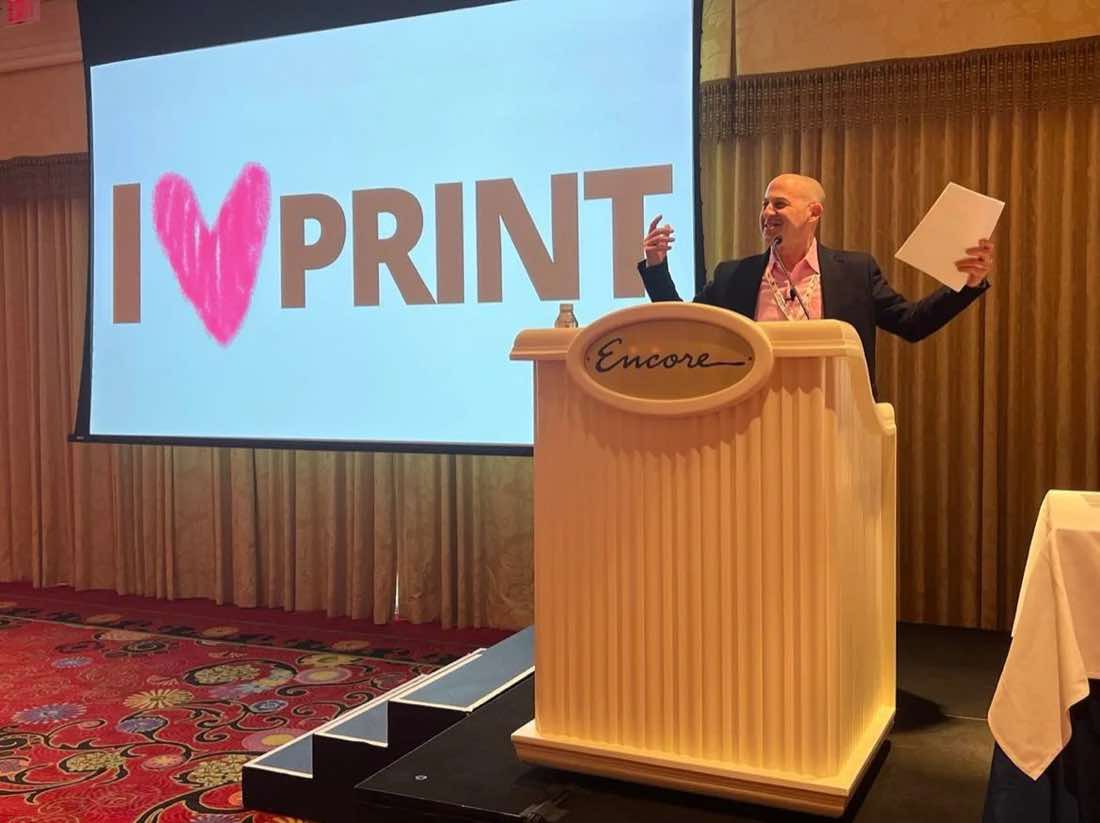 Keynote speaker gesturing at an Encore podium beneath a large I love PRINT slide