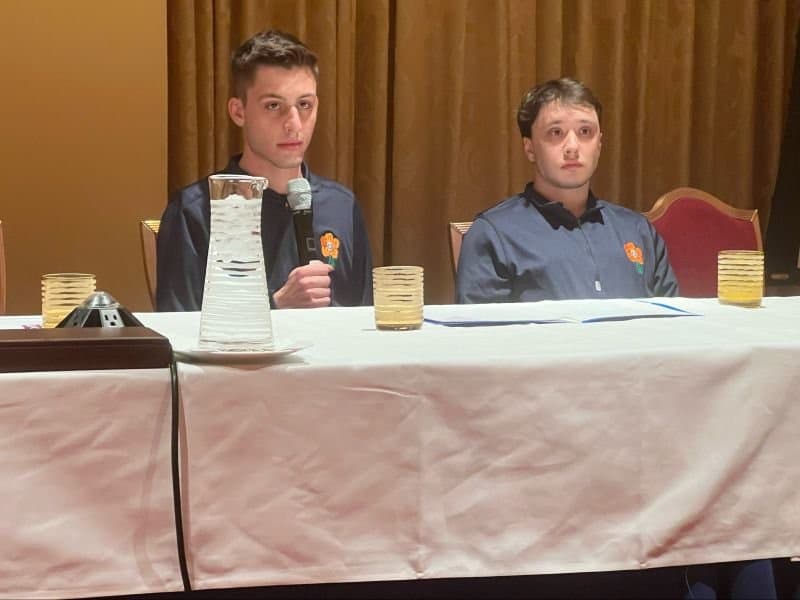 Two speakers seated at a conference head table, one holding a microphone, during a session