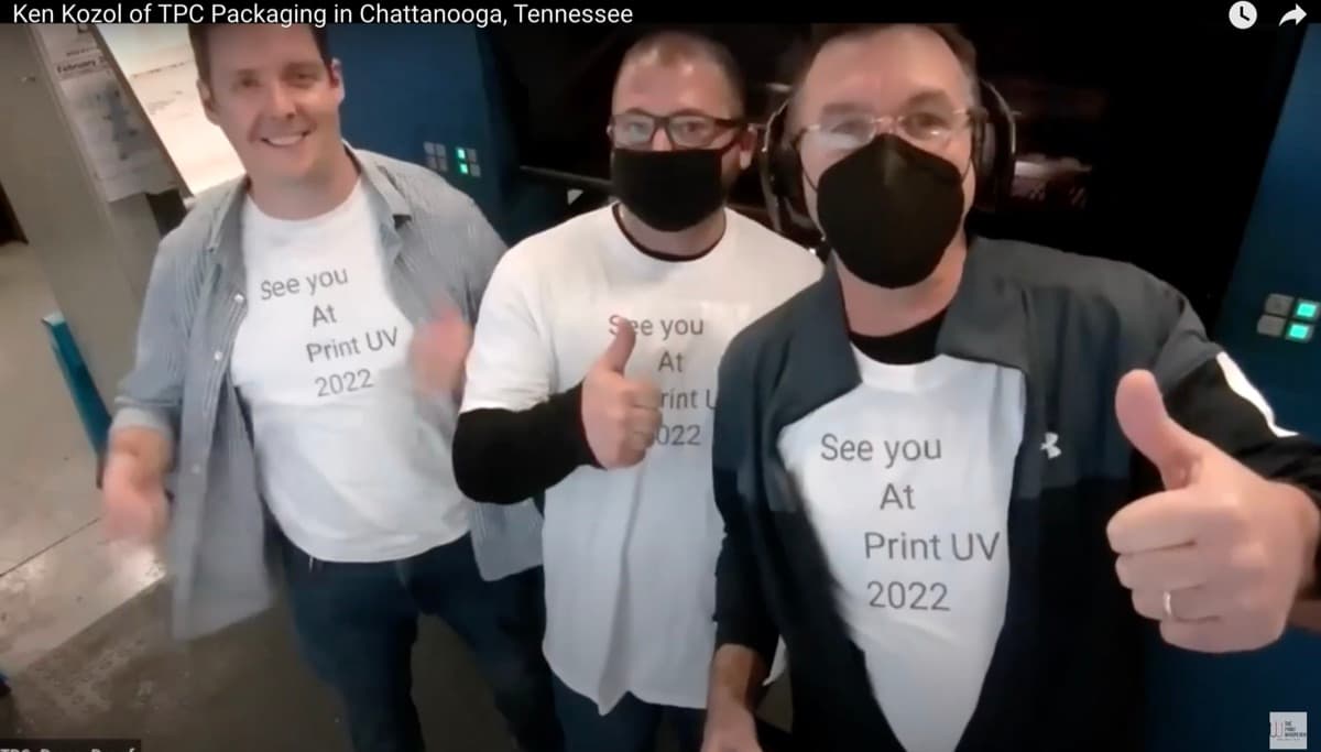 Three TPC Packaging team members in See-you-at-Print-UV-2022 T-shirts giving thumbs-up beside a press
