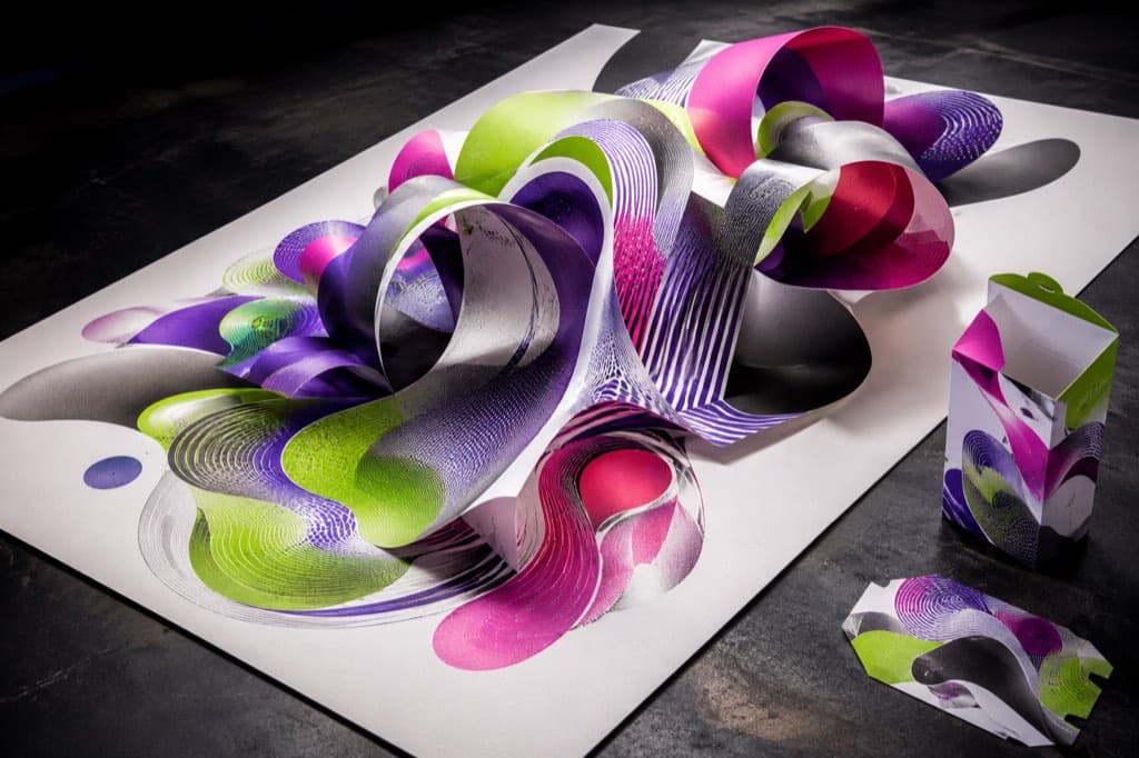 A generative printed pattern rises from a press sheet into dimensional paper ribbons beside a matching carton